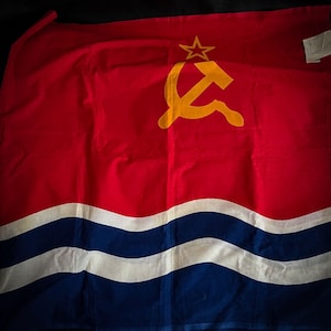 May include: A red flag with a yellow hammer and sickle and a yellow star. The flag has two white stripes and two blue stripes.
