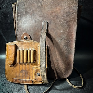 May include: A brown leather satchel with a smaller brown leather pouch attached. The pouch has multiple slots for holding items. The satchel has a long strap for carrying it over the shoulder.
