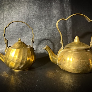 Puede incluir: Dos teteras de latón, una con un diseño acanalado y la otra con una superficie lisa, se encuentran sobre un fondo oscuro.