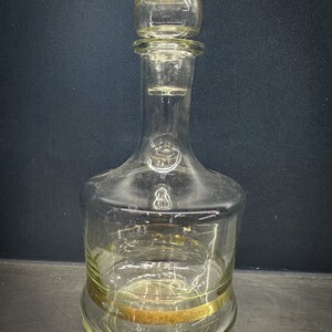 May include: A clear glass decanter with a gold band around the bottom. The decanter has a round, bulbous body and a tall, slender neck.