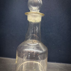 Puede incluir: Una botella de vidrio transparente con un tapón redondo. La botella tiene una base ancha y un cuello estrecho.