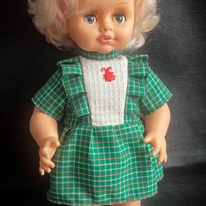 Könnte beinhalten: Vintage Puppe mit blondem, lockigem Haar und blauen Augen. Die Puppe trägt ein grünes kariertes Kleid mit weißem Lätzchen und rotem Stickmuster. Weiße Schuhe vervollständigen das Outfit. Klassisches Spielzeug.