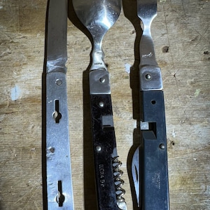 Può includere: Un set di tre utensili da campeggio vintage, tra cui un coltello, un cucchiaio e una forchetta. Gli utensili sono in metallo e hanno manici in plastica nera. Il coltello ha un'apertura a forma di buco della serratura sul manico. Il cucchiaio ha un cavatappi sul manico. La forchetta ha una piccola lama sul manico.