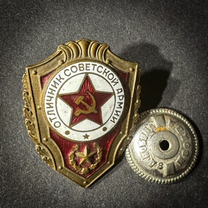 May include: A gold-colored metal badge with a red star and hammer and sickle design. The badge reads "Отличник Советской Армии" which translates to "Excellent Soviet Army".