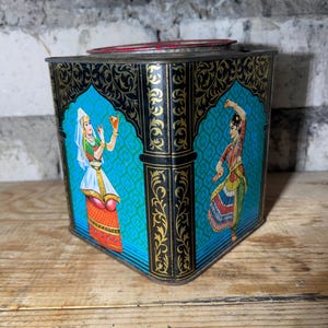May include: Vintage metal tin with a black and gold ornate design. The tin features illustrations of dancing figures in traditional attire against a turquoise backdrop. The lid is red. The tin is sitting on a wooden surface.