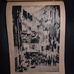 May include: A black and white photograph of a narrow street in a city. The street is lined with buildings and there are clothes hanging from lines strung across the street. There are people walking in the street. The text "TRUOGOLI DI S' BRIGIDA" is printed at the bottom of the image.