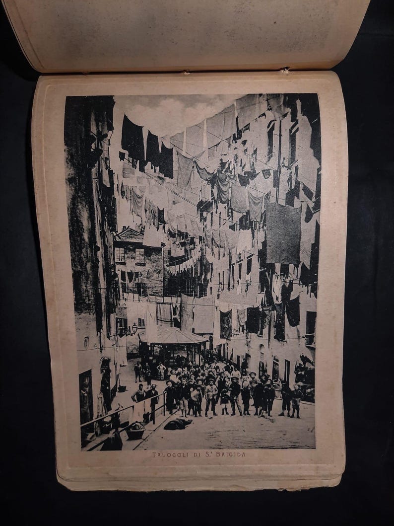 May include: A black and white photograph of a narrow street in a city. The street is lined with buildings and there are clothes hanging from lines strung across the street. There are people walking in the street. The text "TRUOGOLI DI S' BRIGIDA" is printed at the bottom of the image.
