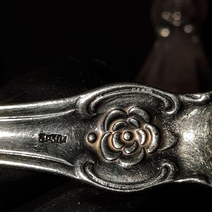 May include: Close-up of a silver utensil with a floral design. The utensil has a detailed rose in the center, with a gold-colored accent. The handle has an ornate, curved design. The text "SP&OK" is visible on the handle.