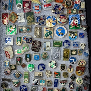 May include: A collection of vintage enamel pins featuring various sports themes, including hockey, soccer, basketball, and track and field. The pins are displayed on a white background and feature a variety of colors, including red, blue, green, yellow, and black.