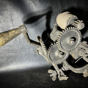 Peut inclure: Un outil vintage à manivelle en fonte avec une poignée en bois et plusieurs engrenages. L'outil a un aspect usé et rouillé et un crochet sur le côté.