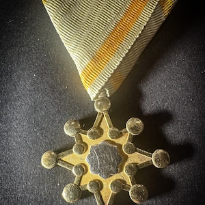 May include: A gold-colored medal with a star-shaped design and a silver center. The medal is attached to a white and yellow ribbon.