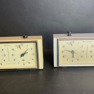 May include: Two vintage chess clocks, one white and one silver, with black hands and red second hands. The clocks have a black border and a white face with black numbers. The white clock says "Sanwa" and "Made in Japan". The silver clock says "Remarkable".