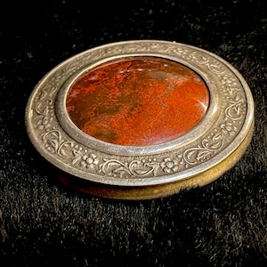 Puede incluir: Un estuche antiguo de plata con un centro de piedra ovalada de color marrón rojizo. El marco de plata presenta intrincados diseños florales y de volutas. El estuche está sobre un fondo oscuro, resaltando sus detalles.
