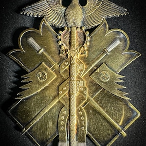 May include: A gold-colored medal with a stylized eagle design, two crossed swords, and a central sword with a tassel. The medal is suspended from a green ribbon.