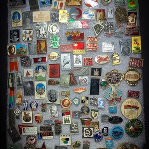 May include: A collection of vintage enamel pins featuring a variety of designs, including buildings, flags, and portraits. The pins are arranged in rows on a gray background.