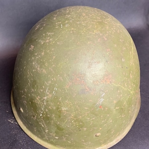 Puede incluir: Un casco militar verde con un acabado desgastado y envejecido. El casco tiene una forma redondeada y una superficie lisa con arañazos y marcas visibles.