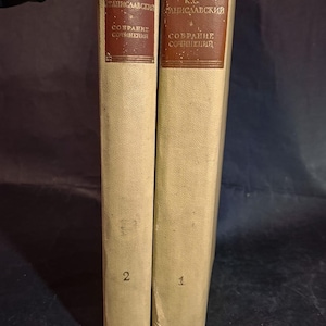 Può includere: Due libri vintage con copertina rigida beige. I dorsi hanno un'etichetta rettangolare marrone scuro con testo in lingua straniera. I libri sono numerati 1 e 2. I libri sono in piedi.