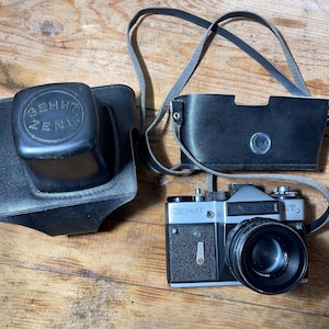 Puede incluir: Una cámara Zenit ET3 vintage con una funda y una tapa de lente de cuero negro. La cámara es plateada y negra, con una lente negra. La tapa de la lente tiene la palabra "ZENIT" impresa. La funda de la cámara tiene una correa para el hombro.