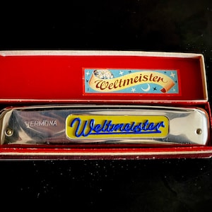 以下が含まれることがあります： 青と黄色の文字で「Weltmeister」と書かれた銀色のハーモニカ。赤い箱に入っています。箱には漫画の画像が付いた小さなラベルが付いています。ハーモニカには「Vermona」と書かれています。