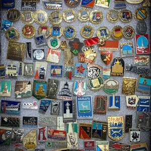 May include: A collection of colorful enamel pins featuring various cityscapes, landmarks, and symbols. The pins are arranged in rows on a gray background. Some of the pins feature text, such as "Moscow", "Samara", and "Ulyanovsk".