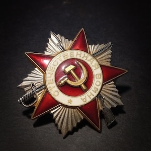 May include: A detailed, close-up view of a vintage Order of the Patriotic War medal. The medal features a red enamel star, a central emblem with a hammer and sickle, and the inscription "ОТЕЧЕСТВЕННАЯ ВОЙНА". The medal is accented with silver details.