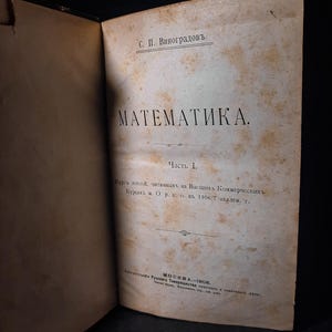 Puede incluir: Un libro antiguo abierto, con páginas amarillentas y envejecidas. El título "MATEMATИKA" se muestra de forma destacada en negrita, con letras cirílicas negras. Se ve texto adicional en ruso, junto con el nombre "С. П. Виноградовъ." La portada del libro es oscura y la estética general es vintage.