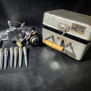 May include: A vintage metal tool kit with a silver-colored hand crank tool and a set of seven silver-colored metal tips. The kit is in a gray metal case with a wooden handle and a label that reads "ЗАВОД МЕХАНИЧЕСКИХ ИЗДЕЛИЙ № 22".