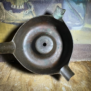 May include: A vintage, dark brown metal candle holder with a circular, indented center and a small hole in the middle. The holder has two curved handles on the sides.
