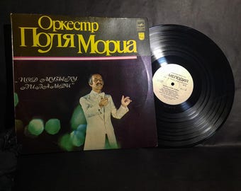 May include: A black vinyl record with a yellow label that says "Melodiya" and a picture of a man in a white suit. The record cover is black with gold lettering that says "Orkestr  Polya Moria" and "pod muzyku  vladeltzu".