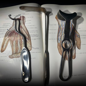 May include: Two surgical instruments, one with a curved handle and a hook at the end, the other with a curved handle and a loop at the end, are laid out on a page of a medical textbook. The textbook page shows a detailed anatomical illustration of a human hand.