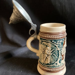 Può includere: Una boccale di birra in ceramica con un coperchio in peltro. La boccale presenta un disegno in rilievo di due uomini in un ambiente boschivo. La boccale è decorata con accenti marroni, verdi e bianchi.