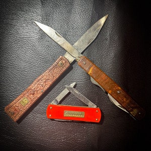 Op de afbeelding: Drie zakmessen met houten en metalen handvatten. De messen zijn gekruist en liggen op een zwart lederen oppervlak. Een mes heeft de tekst "Solingen" op het handvat.