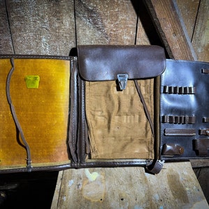 May include: A brown leather case with multiple compartments and a flap closure. The case is open and has a yellow fabric lining.