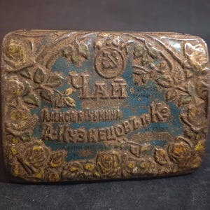 Puede incluir: Una lata de metal vintage con un diseño floral y el texto "Чай" y "Александровский Чай" en escritura cirílica. La lata está oxidada y tiene un fondo azul.