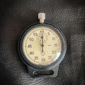 Könnte beinhalten: Eine schwarz-weiße analoge Stoppuhr mit weißem Zifferblatt und schwarzen Zeigern. Die Stoppuhr hat ein schwarzes Kunststoffgehäuse und einen schwarzen Metallring um das Zifferblatt. Das Zifferblatt hat Zahlen von 5 bis 60, und die Worte "MADE IN USSR" sind unten aufgedruckt.