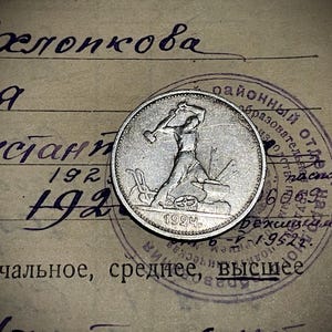 May include: A silver coin featuring a worker with a hammer, dated 1924. The coin is set against a backdrop of aged paper with handwritten text and a circular stamp. The coin's design depicts a figure in action.