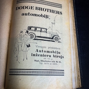 May include: A black and white illustration of a Dodge Brothers automobile with two people standing next to it. The text reads "Dodge Brothers automobili" and "Vienīgais priekšstāvis Automobiļu inženieru birojs (dib. 1910. g.) Rīgā, Elizabetes ielā № 25. Tālr. 30774 un 23578."