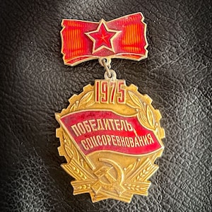 May include: A gold and red metal badge with the text "Победитель Соцсоревнования" (Winner of the Socialist Competition) and the year "1975". The badge features a red star, a hammer and sickle, and wheat stalks.