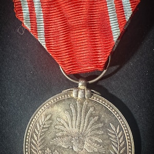 May include: A silver medal with a red and white ribbon. The medal features a stylized bird with a cross and floral designs.