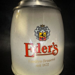 Könnte beinhalten: Ein weißer Keramik-Bierkrug mit einem silbernen Deckel. Der Krug trägt ein rotes und goldenes Logo mit der Aufschrift "Eder's Familien-Brauerei seit 1872".