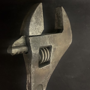 May include: A close-up of a silver adjustable wrench with a worn, textured surface. The wrench has a black handle and a silver adjustment mechanism.