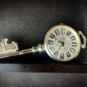 May include: A silver key-shaped clock with a black face and Roman numeral markings. The clock face reads "Slava Jewels". The key is on a black base.