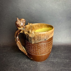 Peut inclure: Une tasse en céramique brune avec une surface texturée et un glaçage jaune à l'intérieur. La tasse a une poignée unique en forme de dragon avec un glaçage brun et noir.