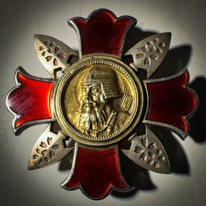 May include: A silver and red cross-shaped medal with a gold medallion in the center. The medallion features a portrait of a man with a beard and a helmet. The medal is decorated with floral designs.
