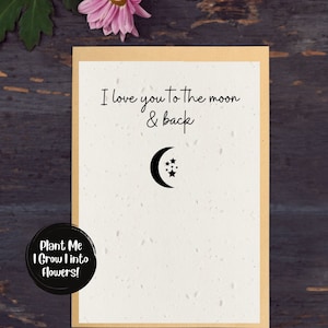 Op de afbeelding: Een crème wenskaart met de tekst "I love you to the moon & back" en een maan en sterren. De kaart ligt op een houten oppervlak met een roze bloem. Een sticker zegt "Plant Me I Grow Into Flowers!"