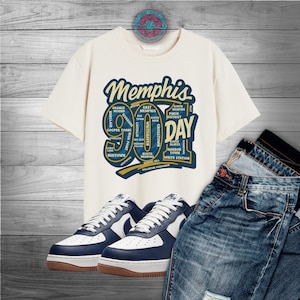 May include: An off-white t-shirt with a Memphis 901 Day graphic, paired with blue jeans and white and navy blue sneakers. The graphic includes the word "Memphis" and various neighborhood names. The sneakers have brown soles.