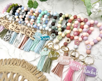 May include: Assortment of colorful silicone beaded bracelets with charms and tassels. The bracelets have different colors and patterns, including floral, marble, and solid colors. The charms include wooden circles with text, acrylic circles with names, and small flower charms. The tassels are made of faux leather and come in various colors.