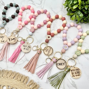 May include: A collection of beaded wrist keychains in various pastel colors, including pink, green, and purple. Each keychain features a tassel and a wooden charm with personalized text. The keychains are designed for holding keys.