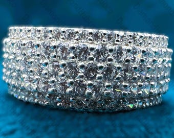 May include: A wide, silver-toned ring densely encrusted with numerous small, clear gemstones. The ring has a multi-layered design, with the gemstones set closely together, creating a sparkling, textured surface. The ring is displayed against a blue background.