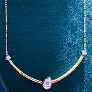 May include: A gold necklace with a curved bar design. It features a pear-shaped, light pink gemstone pendant and two smaller round gemstones. The necklace is set against a teal background.